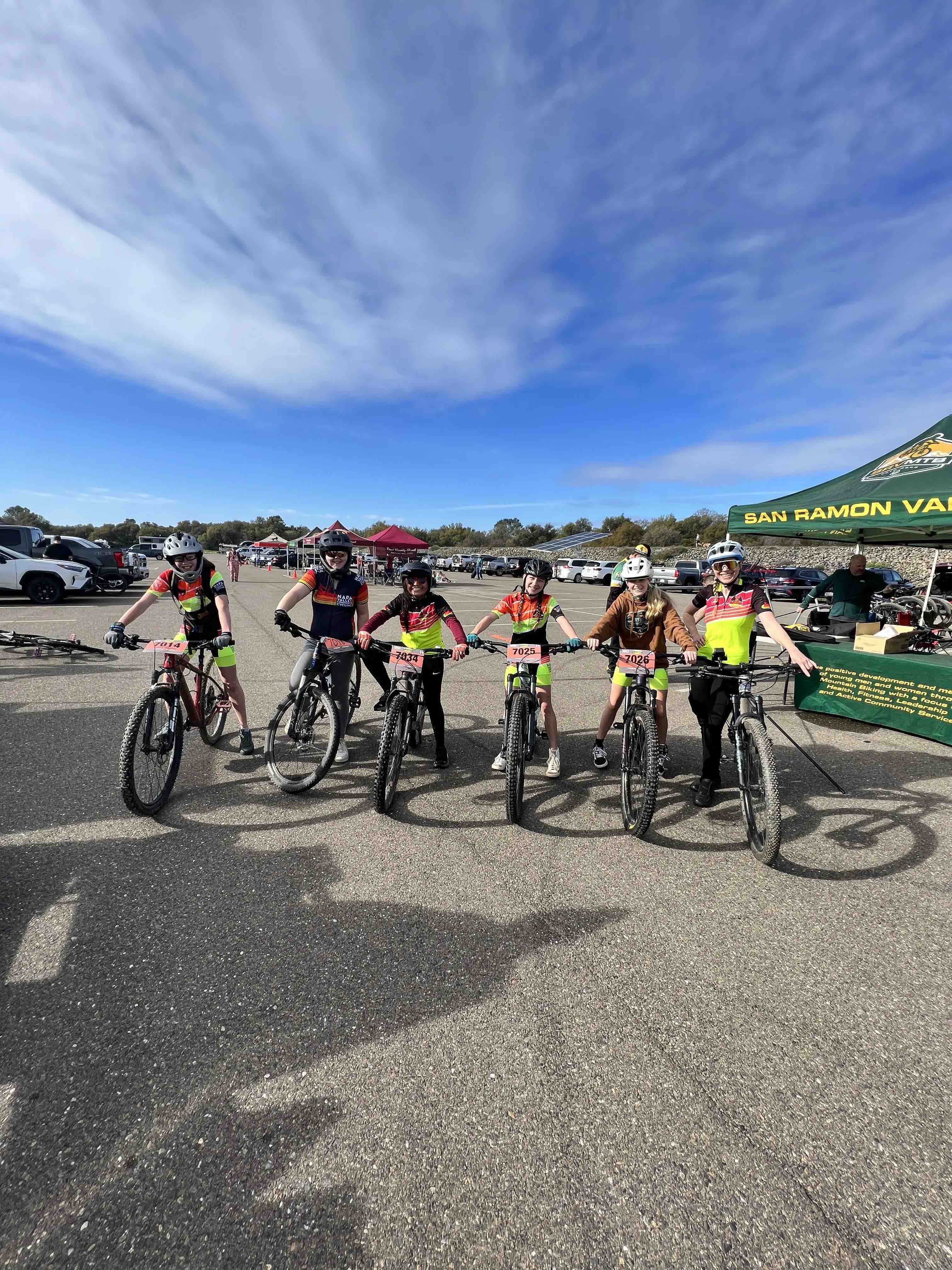 A row of girls on mountain bikes stand beside each other, each wearing their team's uniform and helmet.